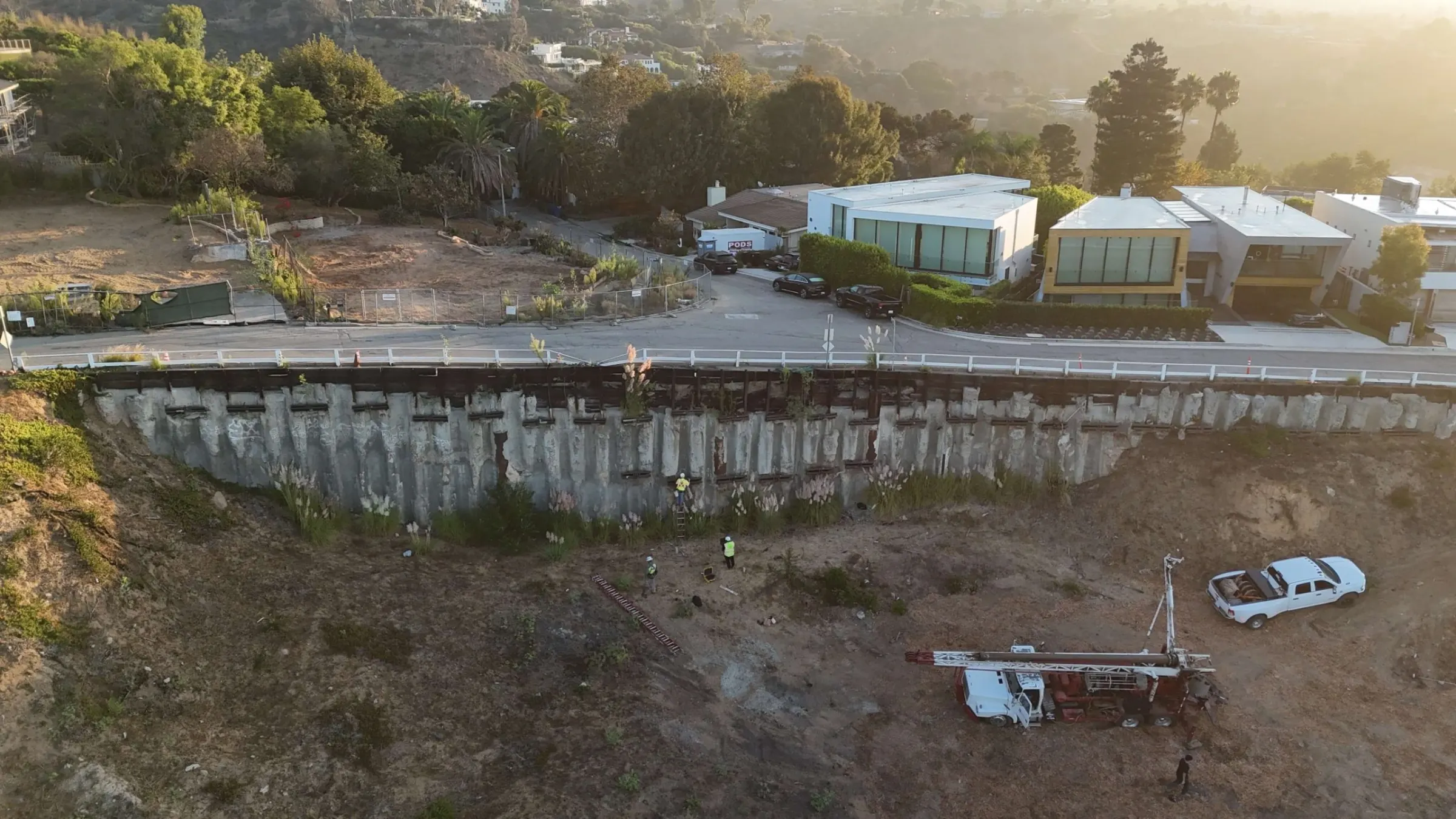 Aerial view of a soil anchor wall below a hillside road, with a drill rig and crew at its base